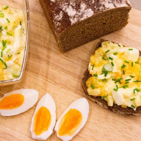 Auf einem Holzbrett liegt eine Scheibe dunkles Roggenvollkornbrot aus der Holzofenbäckerei Ripken, belegt mit cremigem Eiersalat und garniert mit Schnittlauch. Daneben wartet ein ganzer Laib Roggenvollkornbrot neben einer Schüssel Eiersalat und gekochten Eiern in Vierteln mit leuchtendem Eigelb.