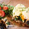 Ein Frühstücksgericht besteht aus pochierten Eiern mit Sauce Hollandaise auf Gersten Vital Gerstenvollkornbrot, belegt mit Spinat und Microgreens. Dazu gibt es geschnittene Champignons und Kirschtomaten auf Rucola.