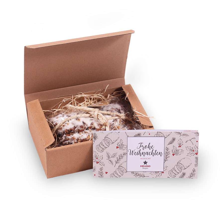 In einem teilweise geöffneten Karton mit strohähnlichem Verpackungsmaterial befindet sich ein mit Puderzucker überzogener Holzofenstollen. Vorne liegt eine festliche Karte, ideal für Firmengeschenke zu Weihnachten.